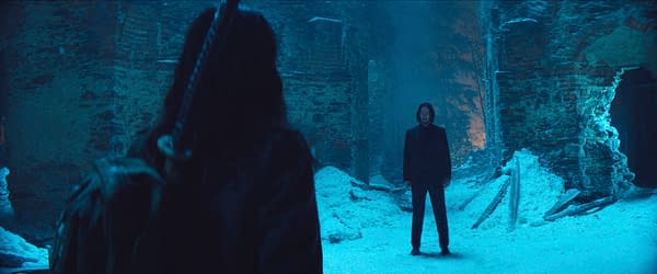 In a snowy, dimly lit setting, Keanu Reeves as John Wick stands face-to-face with an unseen character, creating a tense atmosphere in the film "Ballerina." The abandoned surroundings include partially collapsed walls and snow-covered ground.