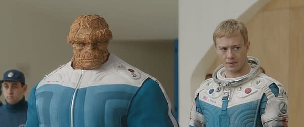 Ebon Moss-Bachrach as Ben Grimm/The Thing stands next to Joseph Quinn as Johnny Storm/Human Torch, both wearing futuristic suits in a scene from 'The Fantastic Four: First Steps.' The background features a modern interior.