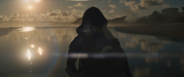A character named Dek, portrayed by Dimitrius Schuster-Koloamatangi, stands on a beach at sunset in the film 'Predator: Badlands.' The character wears a dark hood and a mask, blending into the atmospheric landscape.