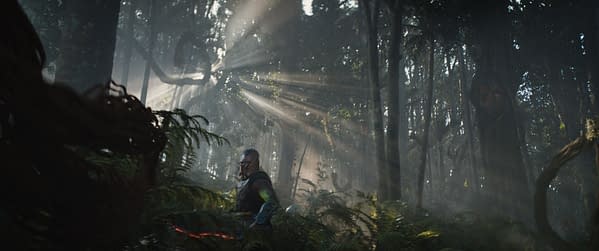 A scene from 'Predator: Badlands' featuring a character named Dek navigating through a dense, misty forest filled with ferns and striking sunlight beams filtering through the trees.