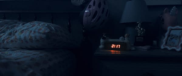 A dimly lit bedroom at night featuring a bedside table with a digital clock displaying 2:17, various small plush toys, a lamp with a horse figurine, and a pink bicycle helmet hanging nearby.
