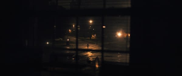 A dark scene viewed through a window, featuring a figure walking down a rain-soaked street illuminated by streetlights, creating a dramatic contrast against the night.