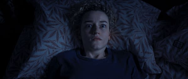 A concerned young woman lies awake in bed, staring wide-eyed at the ceiling, with patterned pillows surrounding her. The scene is dimly lit, suggesting a tense or anxious atmosphere.