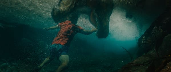A dramatic underwater scene from 'Jurassic World Rebirth,' featuring a person in a red shirt swimming away from a large dinosaur in murky water, emphasizing the tension of the encounter.