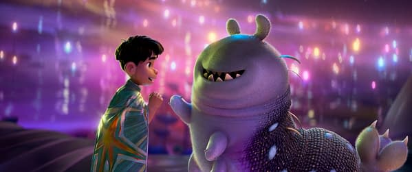 An animation from Disney and Pixar's 'Elio' featuring a young boy in a colorful cloak engaged in a friendly interaction with a whimsical, large alien creature. The background is filled with vibrant, glowing lights, creating a magical atmosphere.