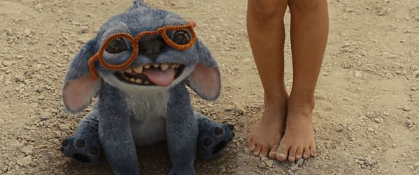 Stitch, a blue alien character from Disney's live-action 'Lilo & Stitch,' is sitting on the ground, wearing orange glasses and playfully sticking out his tongue alongside a child's bare feet.
