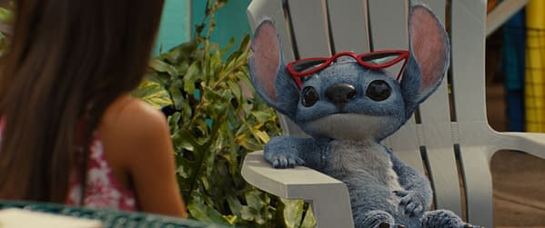 In Disney's live-action Lilo & Stitch, Stitch, the blue alien character, wears red sunglasses and relaxes in a white lawn chair, facing Lilo, who is partially visible in the background surrounded by greenery.