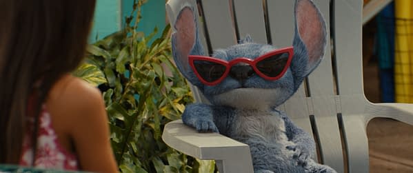 Stitch, a blue alien character from Disney's Lilo & Stitch, sits casually in a chair wearing oversized red sunglasses, while a girl in a floral dress is partially visible in the foreground.