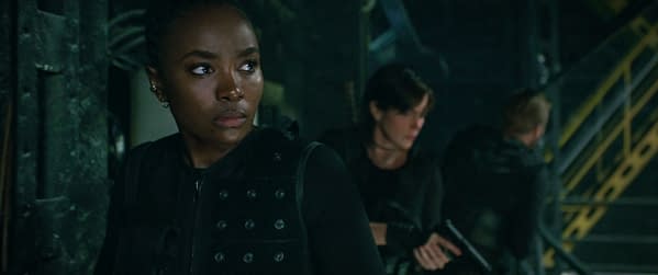 In a scene from The Old Guard 2, KiKi Layne portrays Nile, looking tense and vigilant, while Charlize Theron as Andy is partially visible in the background, holding a weapon. The setting appears dark and industrial, enhancing the intense atmosphere.
