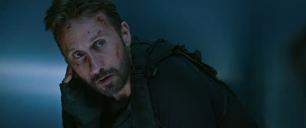 Matthias Schoenaerts as Booker in The Old Guard 2, looking intense with blood smudges on his face, portrayed in a dimly lit setting.