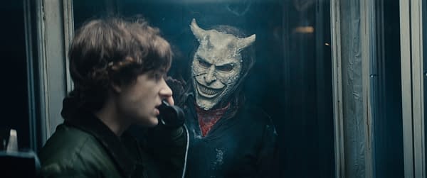 A tense scene from Black Phone 2 features a young boy speaking on a telephone while a sinister figure wearing a devil-like mask looms behind him. This illustration captures the suspenseful atmosphere of the horror film.