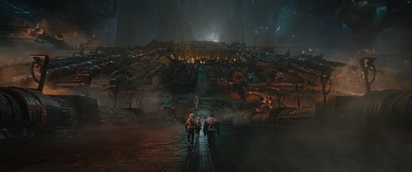 A cinematic scene from 'The Fantastic Four: First Steps,' featuring four characters, including a fiery man and a blue-skinned figure, walking towards a massive, futuristic structure illuminated by eerie lighting, with a misty atmosphere.