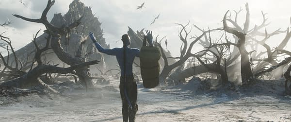 In a desolate landscape from 'Avatar: Fire and Ash', a blue humanoid figure, Quaritch, stands with arms raised, holding a backpack, surrounded by charred trees and a stark, dry terrain.