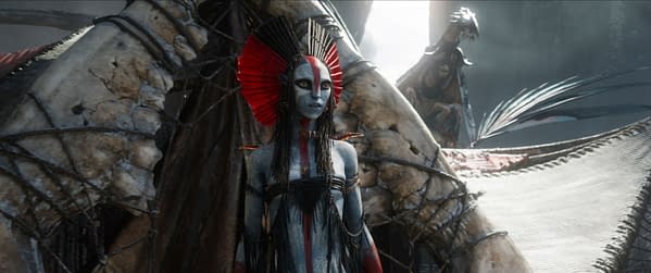 A character named Varang, portrayed by Oona Chaplin, stands in a striking costume with bold red and blue makeup, featuring intricate body markings and elaborate accessories, in a scene from 'Avatar: Fire and Ash'.