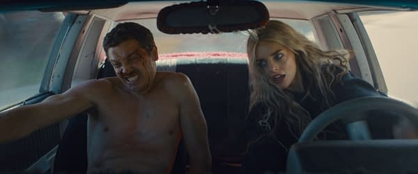 A scene from the film 'Eenie Meanie' features Karl Glusman as John, visibly distressed with a bloodied face, sitting in the driver's seat of a car. Beside him, Samara Weaving as Edie looks intensely focused, both characters conveying a sense of urgency.
