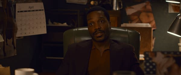 Jermaine Fowler as The Chaperone in the film 'Eenie Meanie,' seated in an office chair. The background features a calendar and various items on the desk.