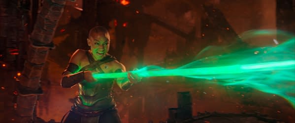A character from Mortal Kombat II wielding a glowing green spear, emitting energy as she prepares to attack. The scene is set in a fiery, chaotic environment.