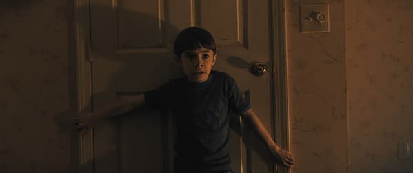 A young boy stands anxiously near a closed door, his expression reflecting fear or surprise. The dimly lit setting features a patterned wallpaper, enhancing the intense atmosphere.