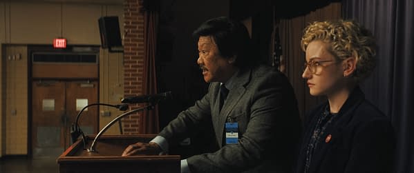 A scene featuring a man speaking at a podium, passionately addressing an audience, while a woman with curly hair and glasses listens intently beside him. The setting appears to be a school auditorium, hinting at a serious discussion, likely related to the themes of the 'Weapons' post.
