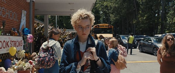 A scene outside a school showing a young woman with curly hair and glasses, looking somber while holding a bag. The background features students and a yellow school bus, with a memorial display reading "LOOK STRONG".