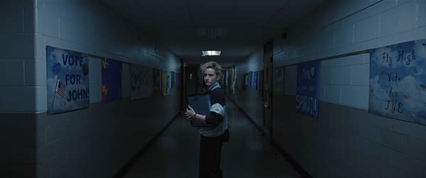A student stands in a dimly lit school hallway, holding a binder, with various campaign posters on the walls advocating for different student candidates. The atmosphere conveys a sense of tension and anticipation.
