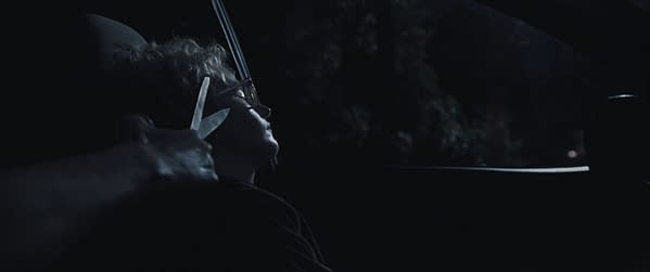 A dramatic still from a scene that appears to take place at night inside a car, featuring a person with glasses sitting in a subdued atmosphere, while a hand holding a pair of scissors is positioned near their face, suggesting tension.