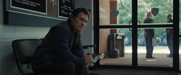A tense scene set in a police station, featuring a man sitting alone on a chair with a posture suggesting contemplation, while two figures converse outside the glass doors. The background includes a police emblem and informational boards on the walls.