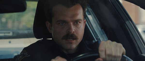 A tense close-up of a police officer driving, showing his focused expression and mustache, with the interior of the police car visible in the background.