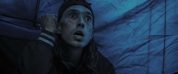 A tense scene showing a young man with long hair and a cap, sitting in a dimly lit environment, his expression conveying fear or anxiety. The background appears to be a blue-toned tent, enhancing the ominous atmosphere.