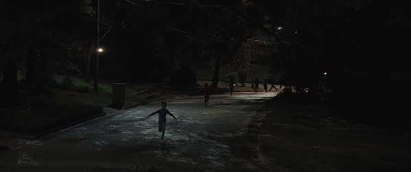 A nighttime scene featuring several children running joyfully down a wet street, illuminated by streetlights. The surrounding area is dark with trees in the background, creating a slightly eerie atmosphere that may relate to a suspenseful moment in a film.