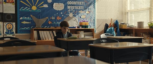 A young boy sits alone at a classroom desk, looking contemplative. The background features a colorful educational bulletin board with marine-themed decorations and motivational phrases.