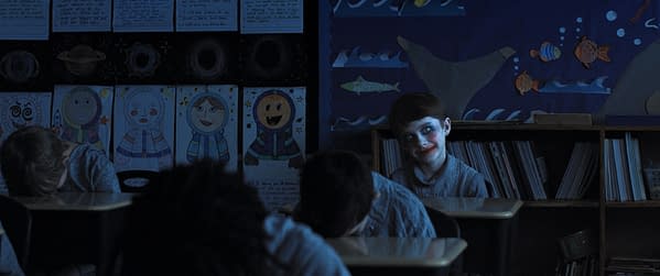 A dimly lit classroom featuring a child with a painted smile and red lipstick, sitting at a desk while other children have their heads down. The background includes colorful drawings of space and aquatic themes on the walls, creating a juxtaposition of innocence and a creepy ambiance.