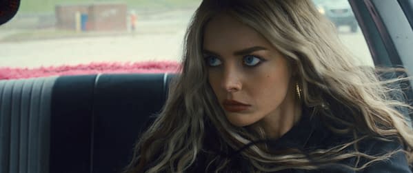 Close-up of a young woman with long, wavy blonde hair and striking blue eyes, who appears intense and focused. She features prominently in the film 'EENIE MEANIE' by 20th Century Studios.