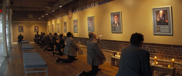 Scene from 'The Naked Gun' showing a group of people kneeling in a hall dedicated to legends, with framed portraits on the wall and candles lit in remembrance.
