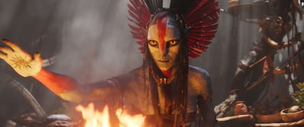 A character named Varang, played by Oona Chaplin, is depicted with a striking painted face and elaborate feather headpiece in a scene from the film 'Avatar: Fire and Ash,' set against a backdrop of fire and smoke.