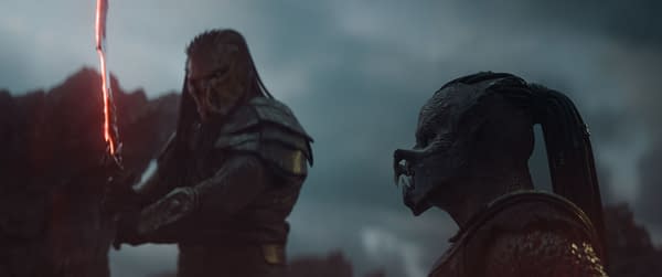 A scene from the film 'Predator: Badlands' features two alien characters, Kwei and Dek, dressed in armor. Kwei holds a glowing sword, while Dek observes amidst a dramatic cloudy background.