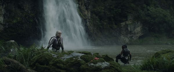 In a scene from the film 'Predator: Badlands,' Thia, portrayed by Elle Fanning, is shown in a dramatic moment wading through water, while Dek, played by Dimitrius Schuster-Koloamatangi, crouches nearby. The backdrop features a cascading waterfall surrounded by lush greenery.