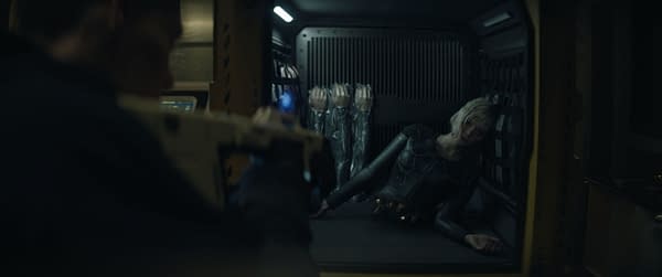 A behind-the-scenes shot from the film 'Predator: Badlands' featuring Elle Fanning as a character in a futuristic setting, lying on a platform behind a transparent barrier, with additional figures in apparent stasis behind her.