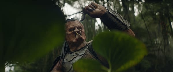 A character from 'Predator: Badlands' is depicted in a dense forest setting, holding a weapon and wearing a detailed costume that includes armor and a distinctive face design. The scene captures the essence of the film's suspenseful atmosphere.