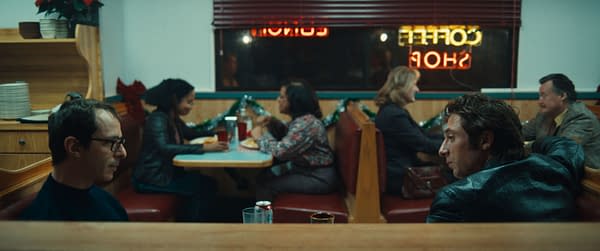 A scene from 'SPRINGSTEEN: DELIVER ME FROM NOWHERE' features two male characters, one in a black turtleneck and glasses, and the other in a leather jacket, seated in a diner. In the background, several people are dining, with festive decorations visible.