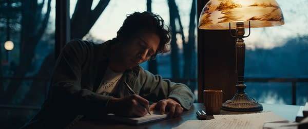 Illustration of Jeremy Allen White portraying Bruce Springsteen, seated at a table and writing in a notebook under a warmly lit lamp, with a window showing an outdoor setting in the background. This image is from 20th Century Studios' film 'SPRINGSTEEN: DELIVER ME FROM NOWHERE'.