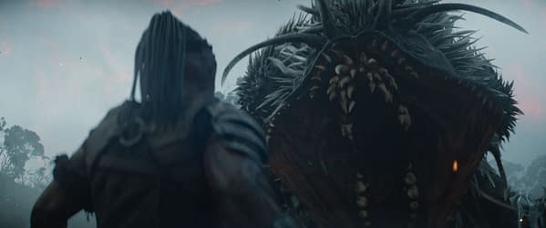 A still from the film "Predator: Badlands" featuring Dimitrius Schuster-Koloamatangi as Dek, facing a large, menacing creature with sharp teeth and spiny features, set against a foggy landscape.