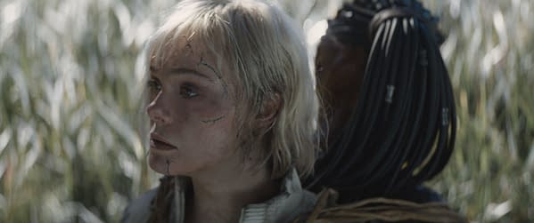 In a scene from 'Predator: Badlands', Elle Fanning portrays Thia, showing signs of distress with scratches on her face, while Dimitrius Schuster-Koloamatangi as Dek is positioned behind her, blending into a natural setting.