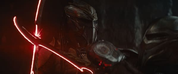 A dramatic scene from 'Predator: Badlands' featuring a character in a detailed armored helmet and fierce expression, wielding a glowing weapon with a red light effect. The atmosphere is dark and intense, capturing the essence of the 20th Century Studios film.