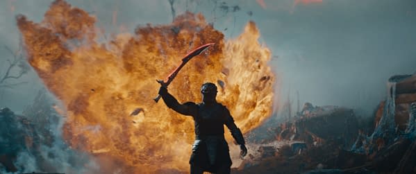A dramatic scene from the film 'Predator: Badlands', featuring a warrior holding a distinctive red sword against a backdrop of flames and destruction.