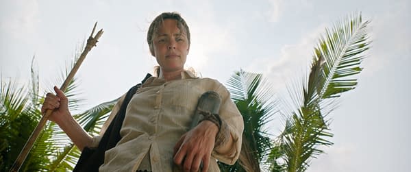 A woman, portrayed by Rachel McAdams as Linda Liddle, stands confidently with a makeshift spear in a tropical setting. Lush palm leaves frame the background under a clear sky.