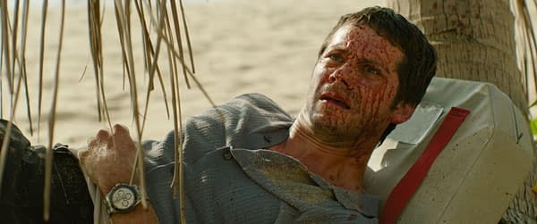 A scene from the film 'SEND HELP' featuring Dylan O'Brien as Bradley Preston, lying on the ground covered in sand and blood, with a distressed expression. The background suggests a beach setting.