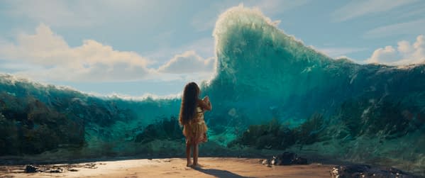 Moana stands on a beach, looking out towards a large, crashing wave in the ocean. The scene is from Disney's live-action adaptation of Moana, highlighting the character's connection to the sea.