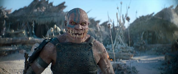 A menacing creature with a toothy grin and intense yellow eyes stands in a desolate landscape, wearing armor and showing detailed facial markings, amidst a backdrop of ruined structures.