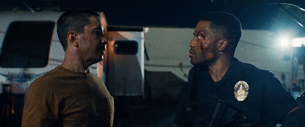 A tense scene from 'Night Patrol' featuring two men facing each other, one dressed in a police uniform and the other in a brown shirt. The officer has visible injuries, including a bruise on his face.
